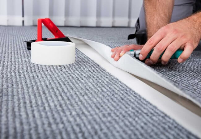 A Close-Up Of A Person Using A Tool To Cut And Install A Seam In Gray Carpet, With Tape Visible.