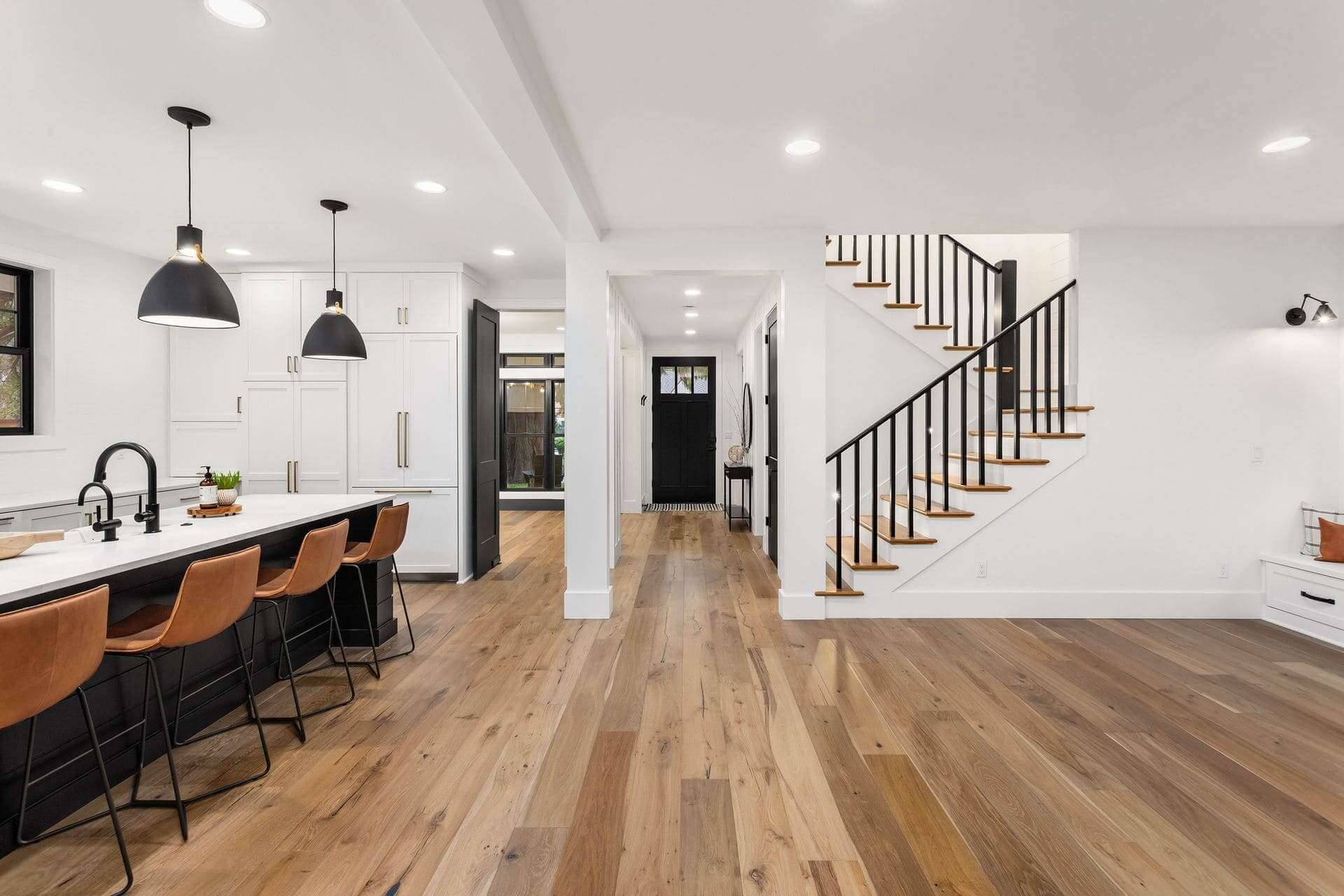 An Open-Concept Home Interior With A Kitchen, Living Area, And Staircase, All Featuring Light-Colored Hardwood Floors.