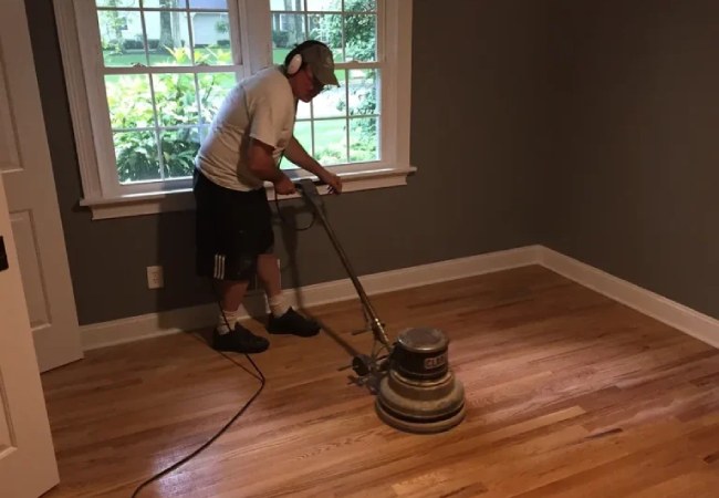 A Man Using A Floor Sanding Machine To Refinish A Hardwood Floor In A Residential Room With Gray Walls.