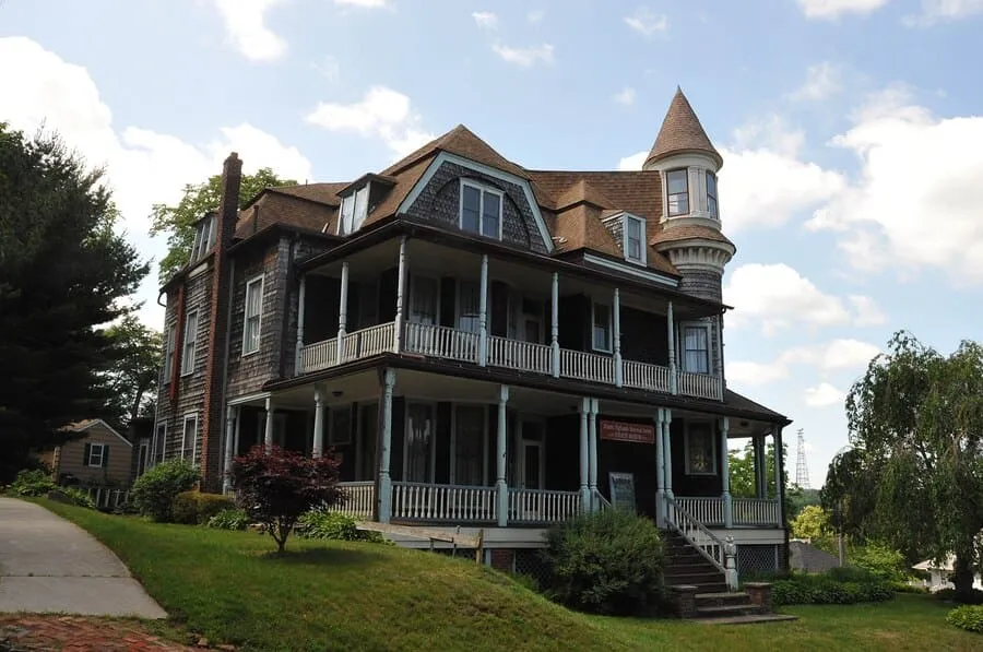A Large, Historic Victorian-Style House With A Prominent Turret, A Wrap-Around Porch, And Dormer Windows, Surrounded By A Green Lawn And Trees.