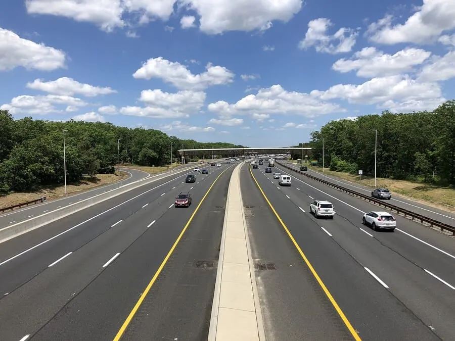 A Wide-Angle View Of A Busy, Multi-Lane Highway With Cars Driving In Both Directions Under A Bright Blue Sky With Scattered Clouds.