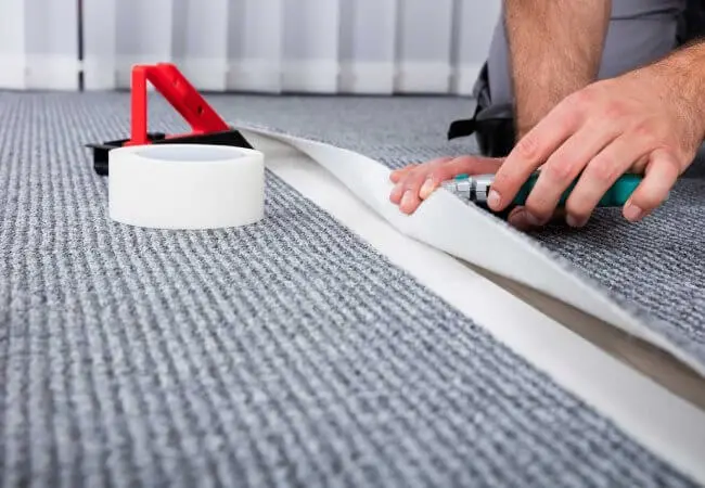 A Close-Up Of A Person Using A Tool To Cut And Install A Seam In Gray Carpet, With Tape Visible.