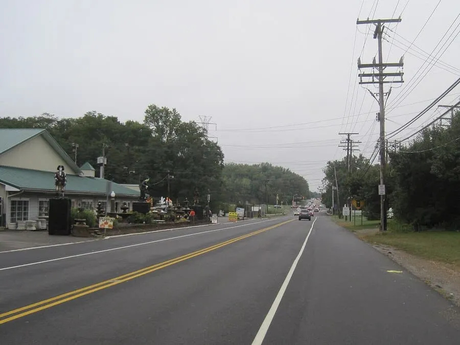 A View Down A Two-Lane Road Lined With Tall Utility Poles And Power Lines On A Cloudy Day, With A Small Building And Trees In The Background.