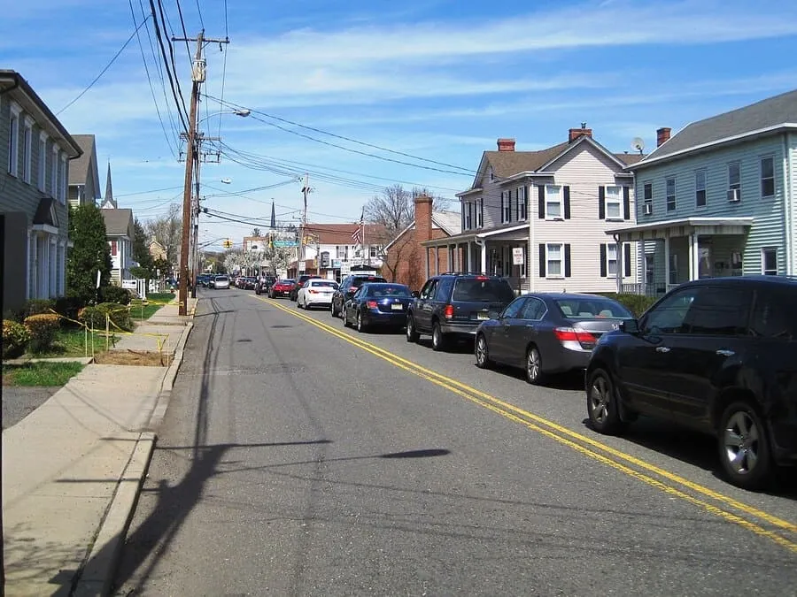 A View Down A Two-Lane Suburban Street With Cars Parked On Both Sides, Lined With Two-Story Houses And Tall Utility Poles Under A Blue Sky With Clouds.