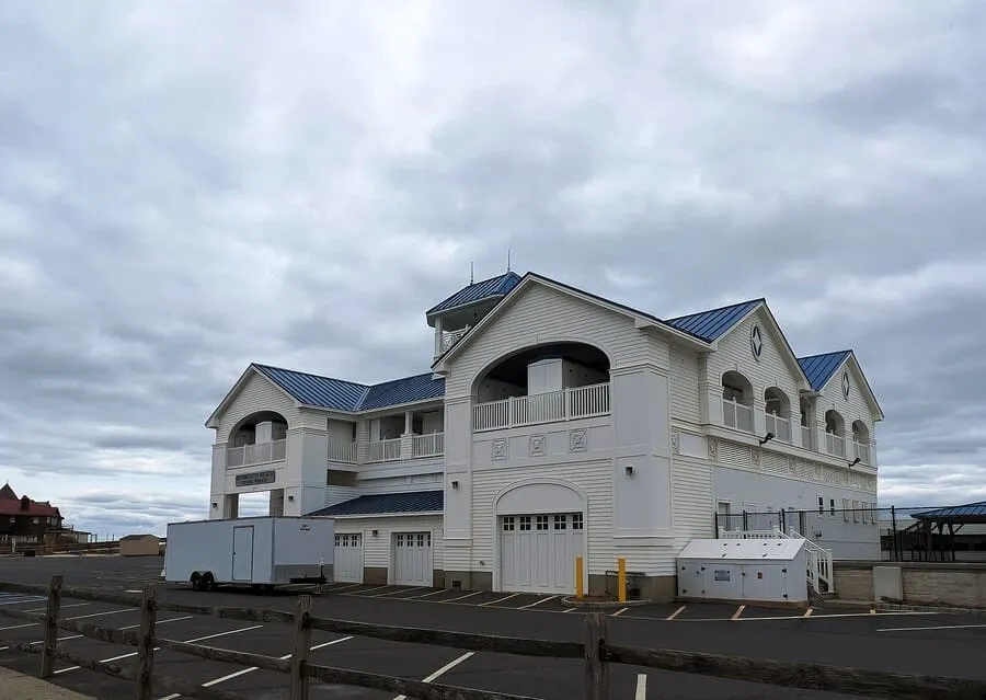 Large, Multi-Level White Building With A Blue Metal Roof And Two Garage Doors Stands On A Paved Lot Next To A Trailer, With A Gray, Cloudy Sky Overhead.