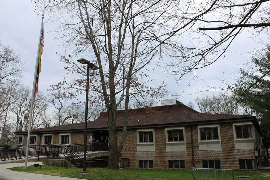 A Two-Story Brick Building With A Brown Roof And A Ramp Leading To The Entrance, With A Flagpole And Bare Tree Branches In The Foreground On A Cloudy Day.